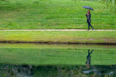 Göz kamaştırıcı bir kadın parkta yürür, zarafeti siyah bir şemsiye ile vurgulanır, yağmurun dünyayı öpmesinden önceki kasvetin ortasında..