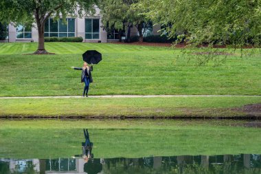 Göz kamaştırıcı bir kadın parkta yürür, zarafeti siyah bir şemsiye ile vurgulanır, yağmurun dünyayı öpmesinden önceki kasvetin ortasında..