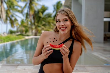 Havuz kenarında ışıl ışıl, genç bir güzellik tatil saadetiyle güneşleniyor. Neşeli bir şekilde karpuzu yiyip bitirirken gülüşü ışıldıyor. Karayipler 'de büyüleyici bir kadın çarpıcı bir sahilde poz veriyor.