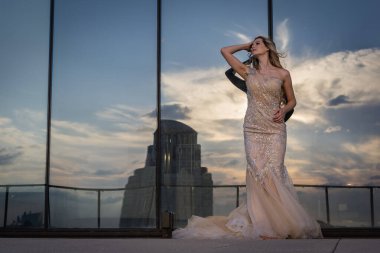 Güneşin altında parlayan güzel bir sarışın manken çarpıcı bir gelinlikle dışarıda poz veriyor. Arkasındaki aynalı bina ışığı yansıtıyor, ışığını arttırıyor ve büyüleyici bir sahne yaratıyor..