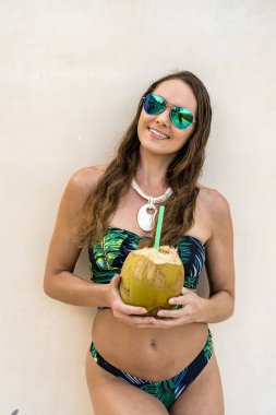 Çarpıcı bir model, Karayip bayramında, cızırtılı bir bikini giymiş, okyanusun kenarında güneşlenen hindistan cevizi içeceğinin tadını çıkarıyor..