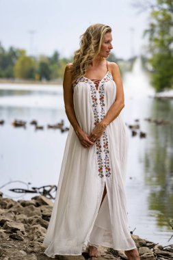 Göz kamaştırıcı sarışın bir model açık beyaz bir elbise giyerek yaz güneşinin tadını çıkarıyor. Huzurlu bir gölün yanında, yumuşak güneş ışığı onun üzerinde dans ediyor, zarafetini ve huzur ortamını arttırıyor..