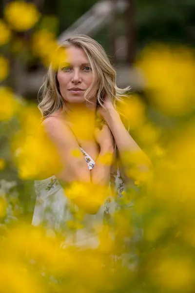 stock image A beautiful blonde model strolls through a field of vibrant yellow flowers, striking poses for portraits. The late summer sun casts a warm glow over the scene, highlighting her grace and natural beauty.