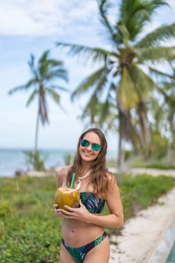 Çarpıcı bir model, Karayip bayramında, cızırtılı bir bikini giymiş, okyanusun kenarında güneşlenen hindistan cevizi içeceğinin tadını çıkarıyor..