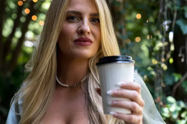 stock image A beautiful blonde woman relaxes in a sun-shaded park, sipping coffee. She smiles, absorbed in her cellphone, enjoying the peaceful surroundings and soft breeze, with trees casting gentle shadows around her.