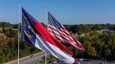 The American flag and North Carolina state flag flutter side by side in the crisp autumn air, vibrant reds, whites, blues, and golds glowing under a clear sky, framed by trees with colorful fall foliage. clipart