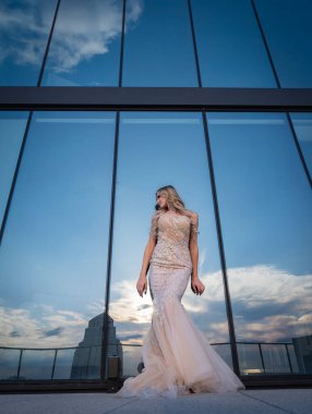 Güneşin altında parlayan güzel bir sarışın manken çarpıcı bir gelinlikle dışarıda poz veriyor. Arkasındaki aynalı bina ışığı yansıtıyor, ışığını arttırıyor ve büyüleyici bir sahne yaratıyor..