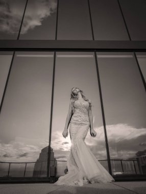 Güneşin altında parlayan güzel bir sarışın manken çarpıcı bir gelinlikle dışarıda poz veriyor. Arkasındaki aynalı bina ışığı yansıtıyor, ışığını arttırıyor ve büyüleyici bir sahne yaratıyor..