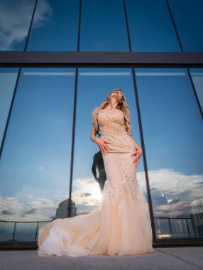 A beautiful blonde model, glowing in the sunlight, poses outdoors in a stunning wedding dress. The mirrored building behind her reflects the light, enhancing her radiance and creating a captivating scene. clipart