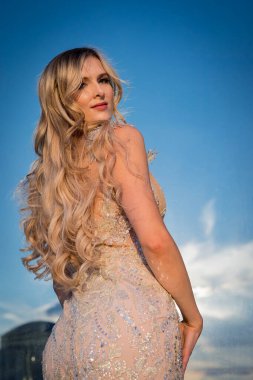 Güneşin altında parlayan güzel bir sarışın manken çarpıcı bir gelinlikle dışarıda poz veriyor. Arkasındaki aynalı bina ışığı yansıtıyor, ışığını arttırıyor ve büyüleyici bir sahne yaratıyor..