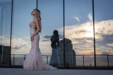 A beautiful blonde model, glowing in the sunlight, poses outdoors in a stunning wedding dress. The mirrored building behind her reflects the light, enhancing her radiance and creating a captivating scene. clipart