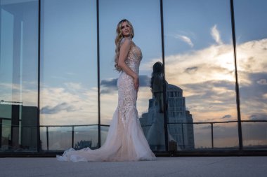 Güneşin altında parlayan güzel bir sarışın manken çarpıcı bir gelinlikle dışarıda poz veriyor. Arkasındaki aynalı bina ışığı yansıtıyor, ışığını arttırıyor ve büyüleyici bir sahne yaratıyor..