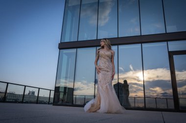 Güneşin altında parlayan güzel bir sarışın manken çarpıcı bir gelinlikle dışarıda poz veriyor. Arkasındaki aynalı bina ışığı yansıtıyor, ışığını arttırıyor ve büyüleyici bir sahne yaratıyor..