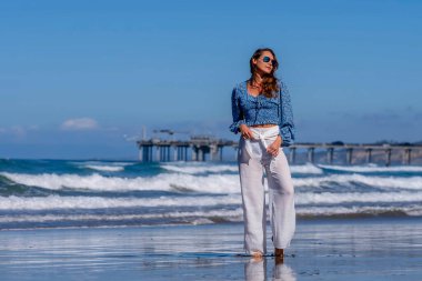 Nefes kesen kadın kumsaldaki mutluluğun tadını çıkarıyor, bir rıhtımın yanında, açık mavi gökyüzünün altında, deniz kıyısında mükemmel bir günün dinginliğini kucaklıyor.