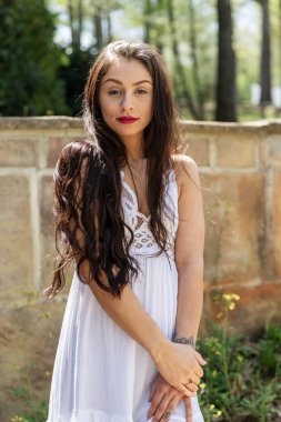 Çekici genç bir esmer, dalgalı beyaz bir bahar elbisesi içinde, saçları dalgalar halinde dalgalanırken dışarıda poz veriyor. Gülümsüyor, uzun ve sert bir kışın ardından baharın yumuşak sıcağında güneşleniyor..