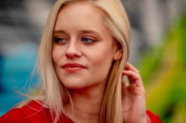 Güzel sarışın bir kadın, şık bahar kıyafetlerini göstererek kameraya neşe içinde poz veriyor. Parlak bir gülümseme ve neşeli tavırlarıyla yaklaşan muhteşem havayı kucaklayarak mutluluk saçıyor..