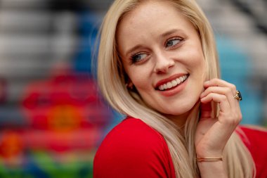 Güzel sarışın bir kadın, şık bahar kıyafetlerini göstererek kameraya neşe içinde poz veriyor. Parlak bir gülümseme ve neşeli tavırlarıyla yaklaşan muhteşem havayı kucaklayarak mutluluk saçıyor..