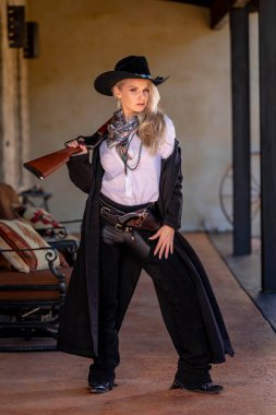 Klasik kovboy kıyafetli, kendine güvenen, güzel, sarışın bir kadın uzun boylu, kalçasında bir tabanca ve omzunda bir tüfekle poz veriyor. Gülüşü enerji ve korkusuz cazibe saçıyor..