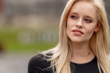 Güzel sarışın bir kadın, şık bahar kıyafetlerini göstererek kameraya neşe içinde poz veriyor. Parlak bir gülümseme ve neşeli tavırlarıyla yaklaşan muhteşem havayı kucaklayarak mutluluk saçıyor..