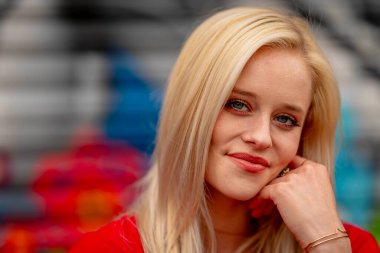 Güzel sarışın bir kadın, şık bahar kıyafetlerini göstererek kameraya neşe içinde poz veriyor. Parlak bir gülümseme ve neşeli tavırlarıyla yaklaşan muhteşem havayı kucaklayarak mutluluk saçıyor..