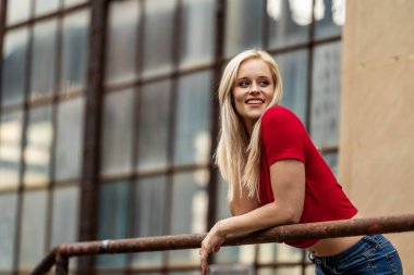 Güzel sarışın bir kadın, şık bahar kıyafetlerini göstererek kameraya neşe içinde poz veriyor. Parlak bir gülümseme ve neşeli tavırlarıyla yaklaşan muhteşem havayı kucaklayarak mutluluk saçıyor..