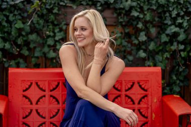 Güzel sarışın bir kadın, şık bahar kıyafetlerini göstererek kameraya neşe içinde poz veriyor. Parlak bir gülümseme ve neşeli tavırlarıyla yaklaşan muhteşem havayı kucaklayarak mutluluk saçıyor..