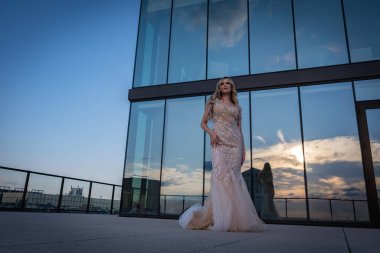 Güneşin altında parlayan güzel bir sarışın manken çarpıcı bir gelinlikle dışarıda poz veriyor. Arkasındaki aynalı bina ışığı yansıtıyor, ışığını arttırıyor ve büyüleyici bir sahne yaratıyor..