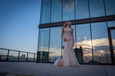 Güneşin altında parlayan güzel bir sarışın manken çarpıcı bir gelinlikle dışarıda poz veriyor. Arkasındaki aynalı bina ışığı yansıtıyor, ışığını arttırıyor ve büyüleyici bir sahne yaratıyor..