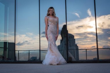 Güneşin altında parlayan güzel bir sarışın manken çarpıcı bir gelinlikle dışarıda poz veriyor. Arkasındaki aynalı bina ışığı yansıtıyor, ışığını arttırıyor ve büyüleyici bir sahne yaratıyor..