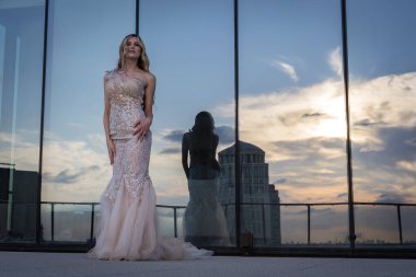 Güneşin altında parlayan güzel bir sarışın manken çarpıcı bir gelinlikle dışarıda poz veriyor. Arkasındaki aynalı bina ışığı yansıtıyor, ışığını arttırıyor ve büyüleyici bir sahne yaratıyor..