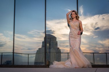 Güneşin altında parlayan güzel bir sarışın manken çarpıcı bir gelinlikle dışarıda poz veriyor. Arkasındaki aynalı bina ışığı yansıtıyor, ışığını arttırıyor ve büyüleyici bir sahne yaratıyor..