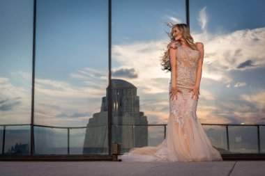 Güneşin altında parlayan güzel bir sarışın manken çarpıcı bir gelinlikle dışarıda poz veriyor. Arkasındaki aynalı bina ışığı yansıtıyor, ışığını arttırıyor ve büyüleyici bir sahne yaratıyor..