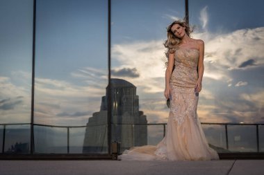 Güneşin altında parlayan güzel bir sarışın manken çarpıcı bir gelinlikle dışarıda poz veriyor. Arkasındaki aynalı bina ışığı yansıtıyor, ışığını arttırıyor ve büyüleyici bir sahne yaratıyor..