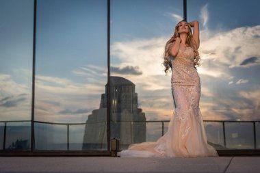 Güneşin altında parlayan güzel bir sarışın manken çarpıcı bir gelinlikle dışarıda poz veriyor. Arkasındaki aynalı bina ışığı yansıtıyor, ışığını arttırıyor ve büyüleyici bir sahne yaratıyor..