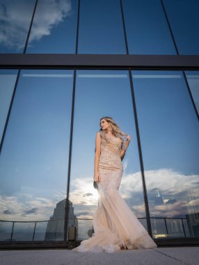 Güneşin altında parlayan güzel bir sarışın manken çarpıcı bir gelinlikle dışarıda poz veriyor. Arkasındaki aynalı bina ışığı yansıtıyor, ışığını arttırıyor ve büyüleyici bir sahne yaratıyor..