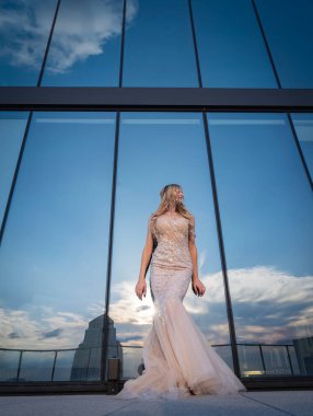 Güneşin altında parlayan güzel bir sarışın manken çarpıcı bir gelinlikle dışarıda poz veriyor. Arkasındaki aynalı bina ışığı yansıtıyor, ışığını arttırıyor ve büyüleyici bir sahne yaratıyor..