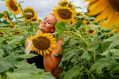 Çekici sarışın bir model canlı bir ayçiçeği tarlasında zarifçe poz veriyor, yaz gününün sıcağında güneşleniyor. Altın çiçekler etrafını sarmış, neşeli ve göz alıcı bir açık hava manzarası yaratıyor..