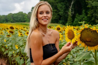 Çekici sarışın bir model canlı bir ayçiçeği tarlasında zarifçe poz veriyor, yaz gününün sıcağında güneşleniyor. Altın çiçekler etrafını sarmış, neşeli ve göz alıcı bir açık hava manzarası yaratıyor..