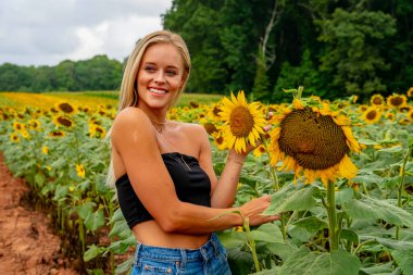 Çekici sarışın bir model canlı bir ayçiçeği tarlasında zarifçe poz veriyor, yaz gününün sıcağında güneşleniyor. Altın çiçekler etrafını sarmış, neşeli ve göz alıcı bir açık hava manzarası yaratıyor..