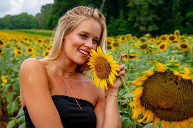 Çekici sarışın bir model canlı bir ayçiçeği tarlasında zarifçe poz veriyor, yaz gününün sıcağında güneşleniyor. Altın çiçekler etrafını sarmış, neşeli ve göz alıcı bir açık hava manzarası yaratıyor..