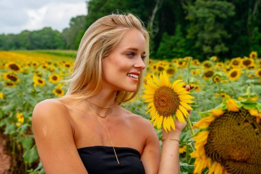 Çekici sarışın bir model canlı bir ayçiçeği tarlasında zarifçe poz veriyor, yaz gününün sıcağında güneşleniyor. Altın çiçekler etrafını sarmış, neşeli ve göz alıcı bir açık hava manzarası yaratıyor..