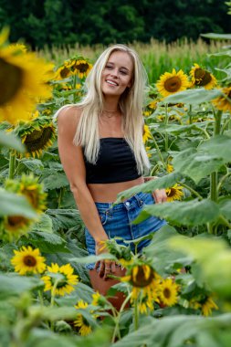 Çekici sarışın bir model canlı bir ayçiçeği tarlasında zarifçe poz veriyor, yaz gününün sıcağında güneşleniyor. Altın çiçekler etrafını sarmış, neşeli ve göz alıcı bir açık hava manzarası yaratıyor..