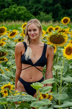 Çekici sarışın bir model canlı bir ayçiçeği tarlasında zarifçe poz veriyor, yaz gününün sıcağında güneşleniyor. Altın çiçekler etrafını sarmış, neşeli ve göz alıcı bir açık hava manzarası yaratıyor..
