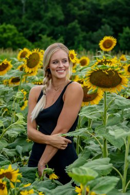 Çekici sarışın bir model canlı bir ayçiçeği tarlasında zarifçe poz veriyor, yaz gününün sıcağında güneşleniyor. Altın çiçekler etrafını sarmış, neşeli ve göz alıcı bir açık hava manzarası yaratıyor..