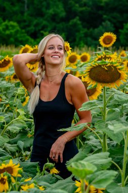 Çekici sarışın bir model canlı bir ayçiçeği tarlasında zarifçe poz veriyor, yaz gününün sıcağında güneşleniyor. Altın çiçekler etrafını sarmış, neşeli ve göz alıcı bir açık hava manzarası yaratıyor..