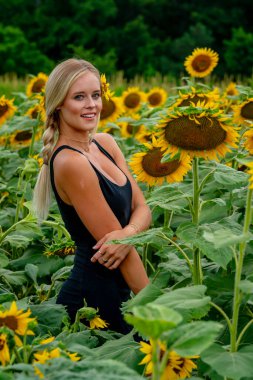 Çekici sarışın bir model canlı bir ayçiçeği tarlasında zarifçe poz veriyor, yaz gününün sıcağında güneşleniyor. Altın çiçekler etrafını sarmış, neşeli ve göz alıcı bir açık hava manzarası yaratıyor..