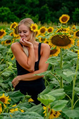 Çekici sarışın bir model canlı bir ayçiçeği tarlasında zarifçe poz veriyor, yaz gününün sıcağında güneşleniyor. Altın çiçekler etrafını sarmış, neşeli ve göz alıcı bir açık hava manzarası yaratıyor..