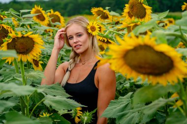Çekici sarışın bir model canlı bir ayçiçeği tarlasında zarifçe poz veriyor, yaz gününün sıcağında güneşleniyor. Altın çiçekler etrafını sarmış, neşeli ve göz alıcı bir açık hava manzarası yaratıyor..