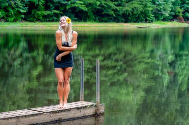 A beautiful blonde woman in a black dress poses gracefully by a serene pond, its calm water reflecting the lush greenery around her. The peaceful setting and vibrant nature evoke a joyful, uplifting mood. clipart