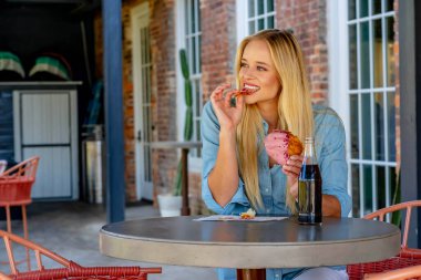 Parlak, genç, şık giyinmiş, pastanın tadını çıkaran ve açık hava alışveriş merkezinde gazoz yudumlayan bir sarışın. O canlı atmosferden ve hafif açık hava esintisinden zevk alırken güneş sıcacık bir parıltı saçar..